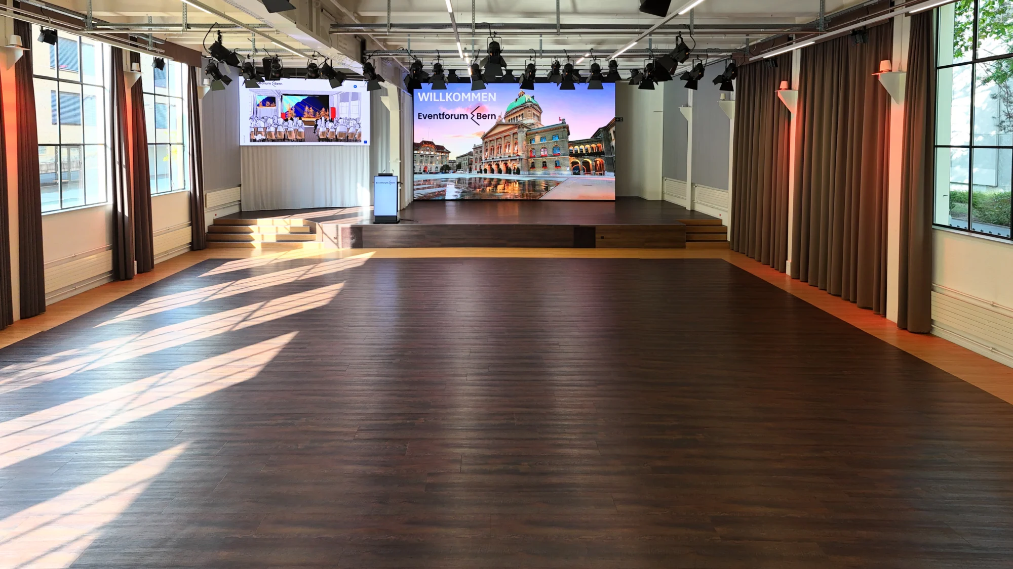 Eventhalle im Eventforum Bern mit freier Saalfläche, Bühne, Rednerpult und zwei LED-Wänden.