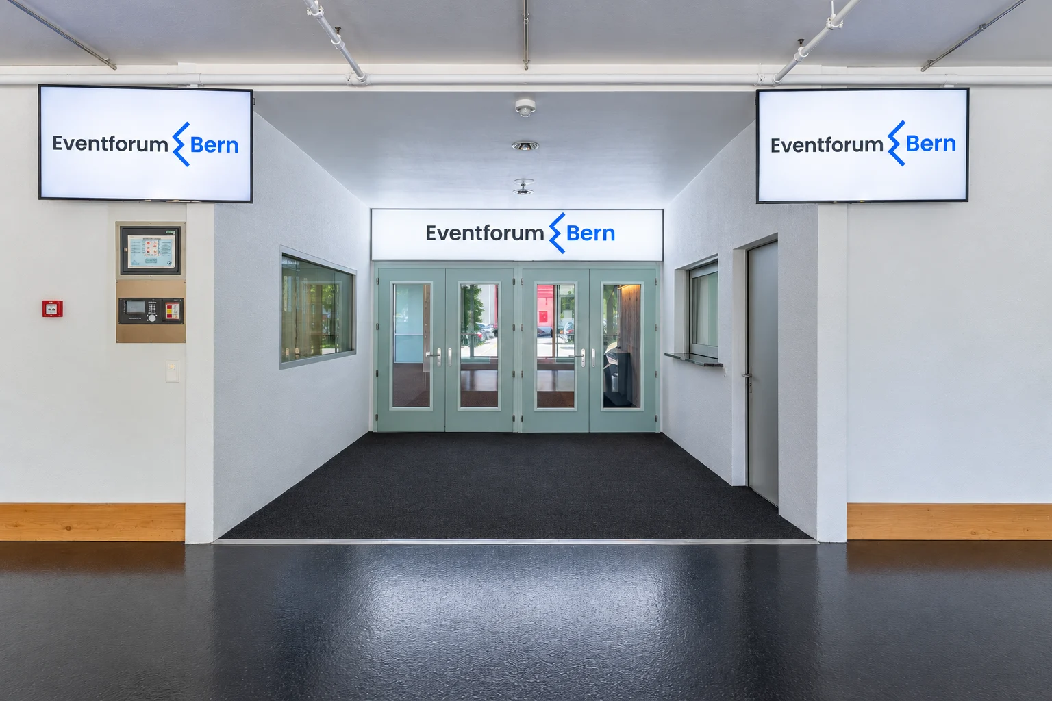 Vorbereich des Eventforum Bern mit aktualisiertem blauem Logo auf den Bildschirmen und über den Eingangstüren.
