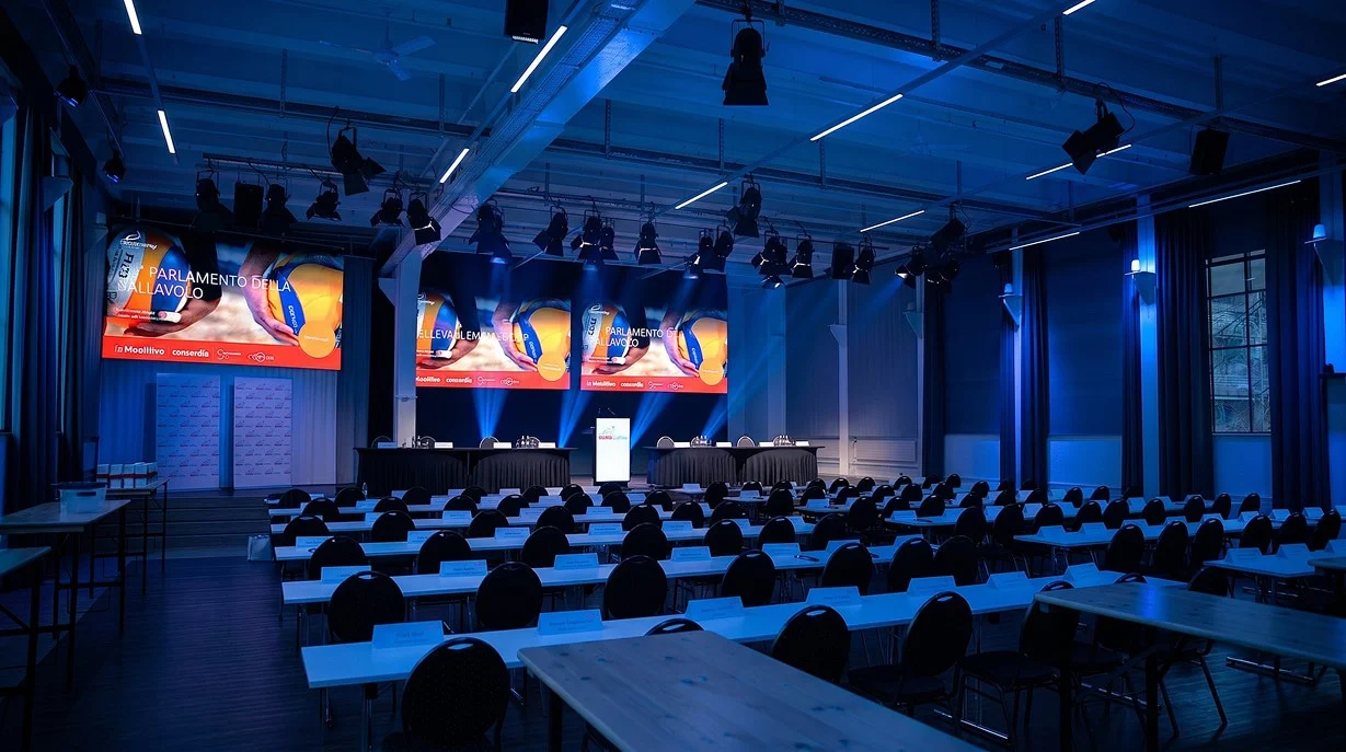 Blau beleuchteter Konferenzsaal mit Bühne, Präsentationsflächen und bestuhlten Tischen im Eventforum Bern.