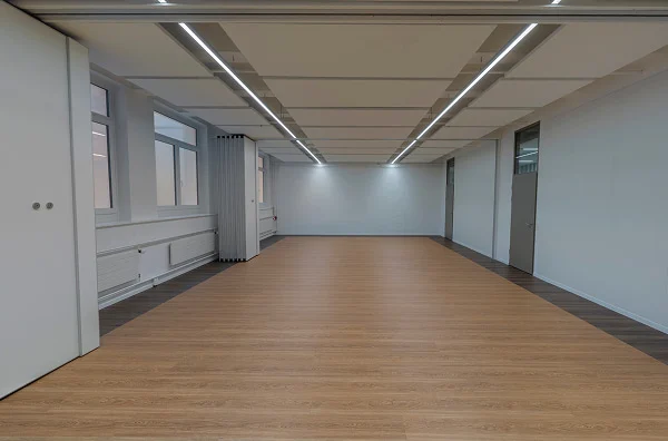 Leerer Workshopraum mit heller Holzfläche, Fenstern und flexiblen Trennwänden.