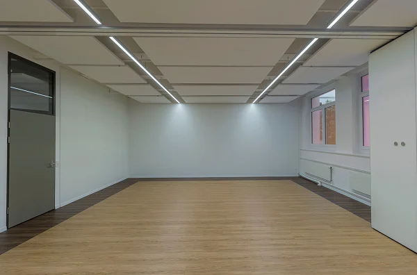Leerer Workshopraum mit heller Holzfläche, Fenstern und moderner Deckenbeleuchtung.