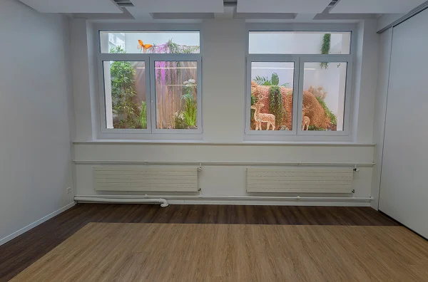 Workshopraum IV mit heller Holzfläche, Fenstern und Blick auf begrünte Innenbereiche.